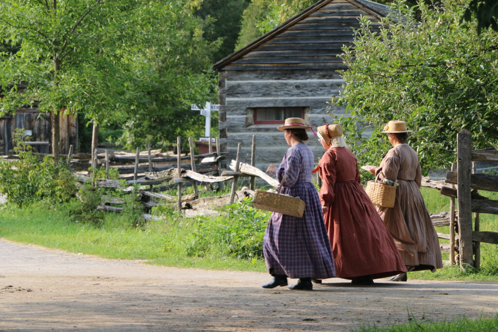 Three Upper Canada Village female interpreters walking away in different coloured dresses in bonnets